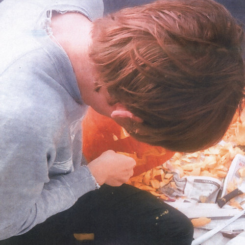 Cover art for Night Times by Fred Thomas. Color photo taken from above shows a person wearing a grey sweatshirt and dark pants carving a pumpkin over laid out newspapers.